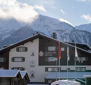 Aussenansicht des Sunstar Hotels Klosters im Chaletstil mit Fahnen vor dem Eingang, dahinter verschneite Berge und Wolken in der Wintersonne