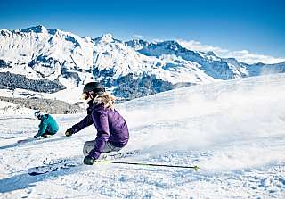Zwei Skifahrer ziehen ihre Kurven im Pulverschnee des Skigebiets Lenzerheide mit beeindruckendem Bergpanorama und strahlend blauem Himmel.