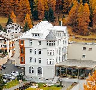 Luftaufnahme des Sunstar Hotels Pontresina mit weisser Fassade und Wintergarten, umgeben von Engadinerhäusern und goldgelben Lärchenwäldern im Herbst
